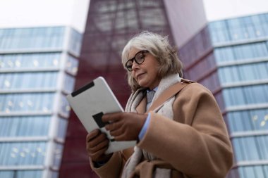 Gri saçlı ve gözlüklü olgun bir kadın, çağdaş cam bir yapının önünde dururken tabletine odaklanıyor. Sahne bir öğleden sonra titreşimini yakalar..