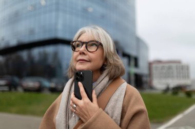 Rahat bir palto ve eşarp giymiş olgun bir kadın dışarıda duruyor, akıllı telefonunu tutarken yukarı bakıyor. Modern şehir manzarası cam binalar ve bulutlu gökyüzü içerir..