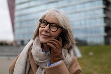 Yaşlı bir kadın, sıcak bir eşarp ve şık eldivenler giyerek cep telefonuyla konuşurken gülümsüyor. Bulutlu bir günde modern şehir mimarisinin ortasında duruyor..