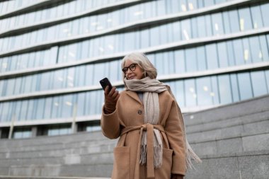 Yaşlı bir kadın, çağdaş bir binanın merdivenlerinde telefonuna bakarken gülümsüyor. Serin hava için kalın giyinmiş, şık eşarbını sergiliyor..