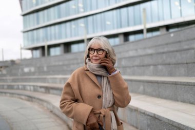 Kahverengi paltolu yaşlı bir kadın şehir binasının önünde dikiliyor, telefonuyla düşünceli bir ifadeyle konuşuyor. Çevre gri ve modern. Havayı serinletiyor..