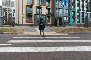Sıcak bir ceket ve şapka giymiş bir çocuk modern bir mahallede yaya geçidinde dikkatle yürüyor. Çevredeki binalar şehir yaşamını gösteriyor.