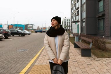 Sıcak bir ceket ve şapka giymiş bir kadın kaldırımda duruyor, soğuk bir günde etrafındakilere derin derin bakıyor. Arka planda modern binalar ve park halindeki arabalar yer alıyor.