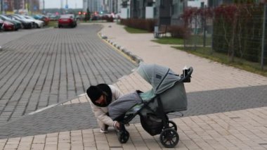 Çağdaş binaların yanındaki geniş kaldırımda bebek arabasını ayarlamak için diz çöken bir kadın görülüyor. Sakin bir gün ve şehir manzarası onu kuşatıyor..