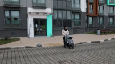 Bir anne bebeğiyle çağdaş bir apartmanın önünde bebek arabasıyla gezer. Sahnede serin bir atmosfer ve iyi döşenmiş bir yol var..