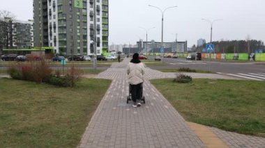 Bir ebeveyn, bir bebek arabasını iterken sakince patikada gezinerek, şehir merkezinde belirlenmiş bir yürüyüş yolu boyunca yürür. Arkadaki binalar yükseliyor..