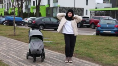 Sıcak giyimli bir kadın, kulaklarını soğuk havadan korurken bebek arabasının yanında duruyor. Gündüz saatlerinde kalabalık bir kentsel alanda modern binalar bir zemin sağlar..