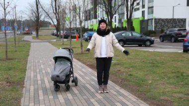 Sıcak giyinmiş bir kadın, şehir merkezinde bir bebek arabasıyla kaldırımda yürüyor. Hava serin görünüyor, çevre de modern binalar ve yeşillikleri gösteriyor..