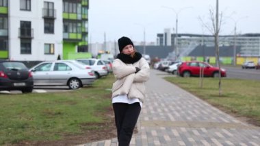 Sıcak, büyük bir ceket ve siyah pantolon giymiş bir kadın serin bir günde kaldırımda yürüyor. Arabalar cadde boyunca park halinde, şehir ortamını vurguluyorlar..