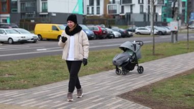 Sıcak giyinmiş bir kadın serin bir günde şehir parkında yürüyor. Neşeli görünüyor ve yakınlarda bir puset park ederken el kol hareketi yapıyor..