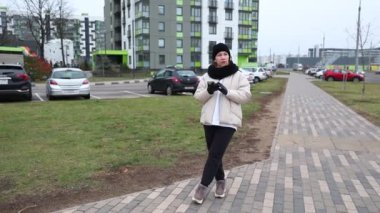 Bir kadın serin hava için sıcak giyinirken kaldırımlı bir patika boyunca yürüyor. Çevresi modern bir apartman kompleksi ve şehir merkezinde park edilmiş arabalarla çevrili..