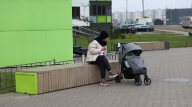 Kışlık ceketli bir kadın bir bankta oturur, yanında bir bebek arabasını izlerken huzur içinde atıştırır. Kentsel çevrede modern binalar yer almaktadır.