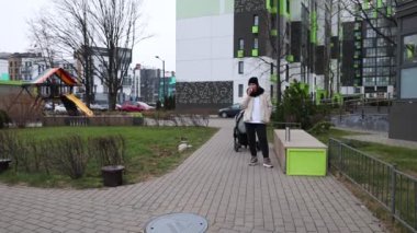 Bir kadın, modern bir parktaki bebek arabasıyla çağdaş binalar ve bulutlu gökyüzünün altında neşeli manzaralarla dolu bir parkta gezintiye çıkıyor..
