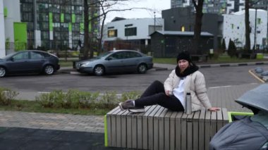 Çağdaş bir şehir parkında şık bir bankta rahatça oturan bir kadın. Etrafında yeşil alanlar, modern binalar ve park edilmiş arabalar var. Atmosfer sakin ve sakin..