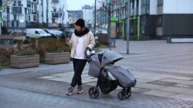 Bir kadın, bebek arabasına rehberlik ederek modern bir mahallede geziniyor. O sakin bir akşam gökyüzü altında akıllı telefonuyla meşgul, günlük hayatı sergiliyor..