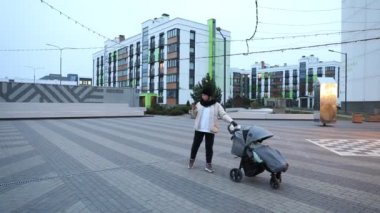 Çağdaş bir meydanda duran bir kadın bebek arabasına eşlik ederken akıllı telefonunu kontrol ediyor. Bölgede modern binalar ve yumuşak akşam ışıkları yer alıyor..