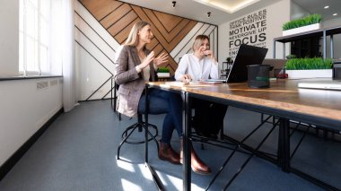 İki kadın şık bir ofiste oturmuş, fikirleri paylaşıyor ve projeleri tartışıyorlar. Parlak güneş ışığı odayı dolduruyor, modern tasarımı ve coşkulu ifadelerini geliştiriyor..