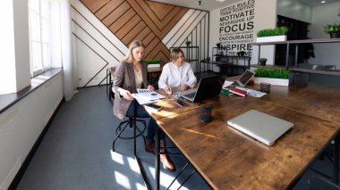 Çağdaş bir ofiste iki kadın şık bir masada birlikte çalışıyor. Raporları ve çizelgeleri gözden geçirirler, parlak, iyi tasarlanmış bir çalışma alanında planlama ve stratejiye odaklanırlar..