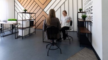İki kadın çağdaş bir ofis ortamında oturmuş sohbet ediyorlar. Mekanda şık mobilyalar ve ilham verici bir duvar tasarımı var. Üretken bir atmosfer yaratıyorlar..