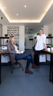 İki kadın rahatlar ve geniş, çağdaş bir ofiste sohbet ederler. Biri sırayla masaya yaslanırken diğeri de sırıtarak ve elinde kahve fincanları tutarak oturuyor. Dekorasyon bitkilerle minimalistik..