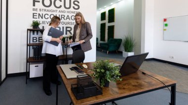 Çağdaş bir ofis ortamında, iki kadın belgeleri incelerken tartışıyorlar. Odaklanmayı ve dürüstlüğü vurgulayan motive edici dekorlarla çevrili..