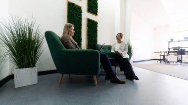 İki kadın rahat yeşil koltuklarda oturuyorlar, şık bir dinlenme odasında düşünceli bir tartışma içindeler, iç mekan bitkileri ve doğal ışıklandırmayla..