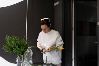 Şık, iyi ışıklandırılmış bir çalışma alanında, odaklanmış bir kadın cam kaplarda taze yeşilliklerle çevriliyken sarı bir laleyi buduyor. Atmosferi geliştirmek için çok dikkatli çalışıyor..