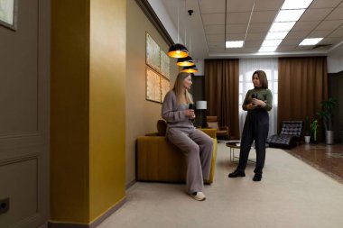 İki kadın, yumuşak ışıklandırma ve çağdaş dekor ile çevrili şık bir salon alanında dostça bir sohbete girişiyor. Biri dinlerken bir bardak tutar..