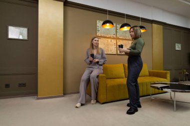 İki kadın oturmuş, modern bir salonda durmuş kahvenin tadını çıkarıyor ve sohbet ediyorlar. Dekorda sarı bir kanepe ve modern bir ışık var, rahat bir atmosfer yaratıyor..