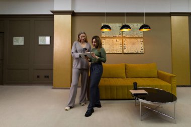 Modern bir oturma odasında yan yana duran iki kadın, mobil bir cihazı paylaşıyorlar. Konforlu alanda sarı bir kanepe, kolye ışıkları ve zarif bir duvar dekoru var..