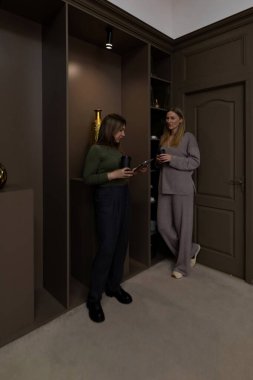 İki kadın şık bir odada durur, biri duvara yaslanır ve bir bardak su içerken diğeri elinde bardakla oturur. Zarif bir dekorun ortasında rahat ve sohbetle meşgul görünüyorlar..