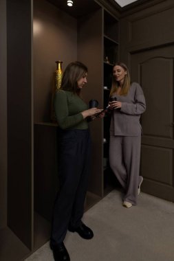 İki kadın şık, tarafsız bir iç mekanda sohbet ediyorlar. Bardaklarını tutarlar ve rahat görünürler. Birlikte geçirdikleri zamanın keyfini çıkarırlar..