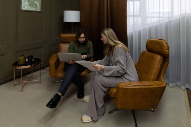 İki kadın şık, modern koltuklarda oturmuş sıcak dekore edilmiş bir oturma odasında belgeleri gözden geçiriyorlar. Hafif ışıklandırma bulutlu bir günde davetkar bir atmosferi güçlendirir..