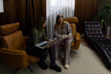 Sıcak ve davetkar bir oturma odasında, iki kadın modern sandalyelerde otururken iş tartışmasına giriyorlar. Biri dizüstü bilgisayar kullanırken, diğeri de belgeleri inceleyerek ortak bir atmosfer yaratıyor..