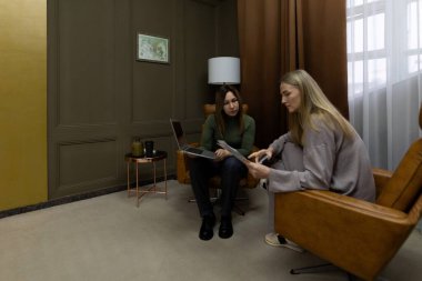 İki kadın şık bir ofiste odaklanmış bir tartışmanın içindeler. Bir kadın tabletinde bir belge gösterirken, diğeri sıcak ışıklandırma ve çağdaş dekor ile çevrili bir şekilde gözlemliyor..