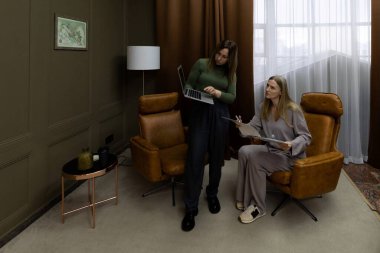 İki kadın zarif ve davetkar bir iç mekanda bir tartışmaya giriştiler. Bir tanesi dizüstü bilgisayar tutarken, diğeri belgeleri inceliyor, şık bir ortamda takım çalışması sergiliyor..