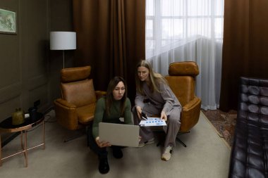 İki kadın modern mobilyaları olan rahat bir yaşam alanında dizüstü bilgisayara odaklanmış durumda. Biri yazarken, diğeri renkli bir palet tutuyor, derin bir tartışmanın içinde..