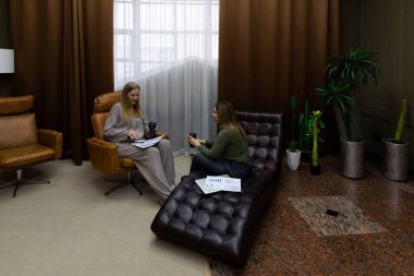 Şık bir salonda, iki kadın rahat bir şezlongda otururken sohbet ederler. Sıcak dekor bitkileri ve yumuşak ışıklandırmayı içeriyor, davetkar bir ortam yaratıyor..