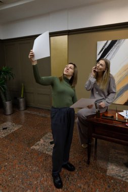 Biri belgeleri havada tutarken, iki çalışan profesyonel belgeler hakkında tartışıyorlar. Dekor, bitkiler ve sanat eserlerinin yer aldığı modern bir ofis alanı..