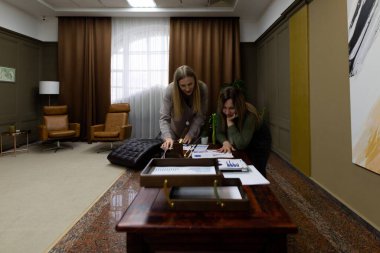 Çağdaş bir ofiste birlikte çalışan iki kadın, ahşap bir masada belge ve çizelgeleri analiz ediyorlar. Konforlu oturma ve dekoratif sanat uzaya profesyonel bir atmosfer ekler..