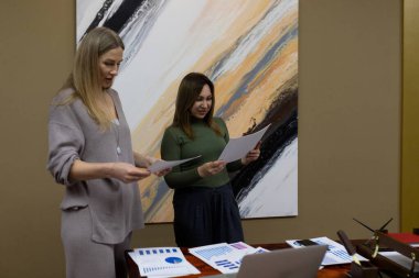 İki kadın bir ofiste birlikte, yazılı raporları ve çizelgeleri inceliyor. Bir kadın gri bir elbise giyer ve diğerinin yeşil bir bluzu vardır. Masada bir dizüstü bilgisayar ve çeşitli belgeler var..