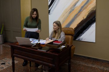 İki kadın modern ahşap bir masada şık bir ofiste birlikte çalışıyorlar. Biri basılı belgeleri incelerken, diğeri de bir dizüstü bilgisayara odaklanarak işbirliği çabalarını destekliyor.