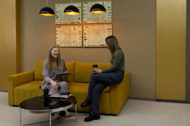 İki kadın şık bir salonda oturup dostça sohbet ediyorlar. Bir kadın tablet tutarken, diğeri sıcak dekorla çevrili bir şekilde içkinin tadını çıkarıyor..