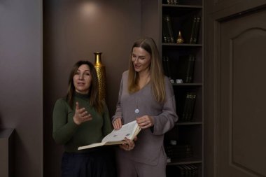 İki kadın modern bir odada durmuş, bir kitabı tartışıyorlar. Bir kadın animasyonlu bir şekilde konuşurken diğeri de şık dekorlar ve rafların arasında kitabı açık tutuyor..