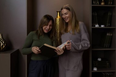 İki kadın, dekoratif eşyalarla süslenmiş şık bir odada birlikte kitap okurken gülümser ve güler. Ortam sıcak ve davetkar. Arkadaşlıklarını sergiliyorlar..