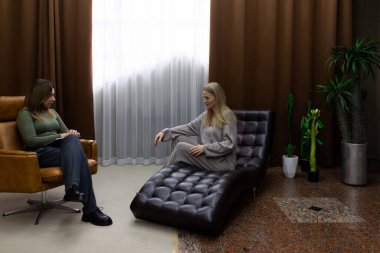 İki kadın, iyi aydınlatılmış bir terapi odasında saksı bitkileriyle dolu düşünceli bir tartışmaya girişiyor. Bir kadın deri bir şezlongda otururken diğeri dikkatle dinliyor..