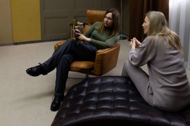 İki kadın, kapalı bir ortamda rahat rahat oturup, hareketli bir sohbete dalarlar. Bir kadın modern bir sandalyede otururken, diğeri lüks bir sedirde uzanır..