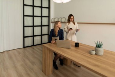 İki kadın, iyi ışıklandırılmış, minimalist bir çalışma alanında konuşurken dijital cihazlar tutuyor. Ahşap bir masa ve basit bir dekor profesyonel atmosferi güçlendirir..