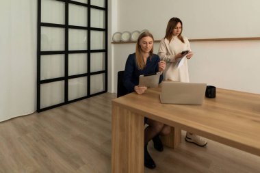 Çağdaş bir ofiste, iki kadın verimli bir çalışma seansı düzenler. Biri ahşap bir masada belgeleri incelerken, diğeri de görevlerine odaklanmış bir tablet kullanıyor..