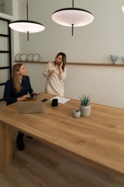 Çağdaş bir ofiste iki kadın ahşap bir masada sohbet ediyorlar. Bir kadın dizüstü bilgisayarla otururken diğeri ayakta telefonla konuşuyor. Ilık ışık sıcak bir ortam ekler..